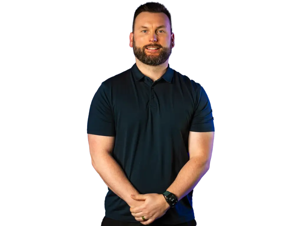 Smiling bearded man in a dark navy polo shirt standing with his hands clasped, against a plain light gray background