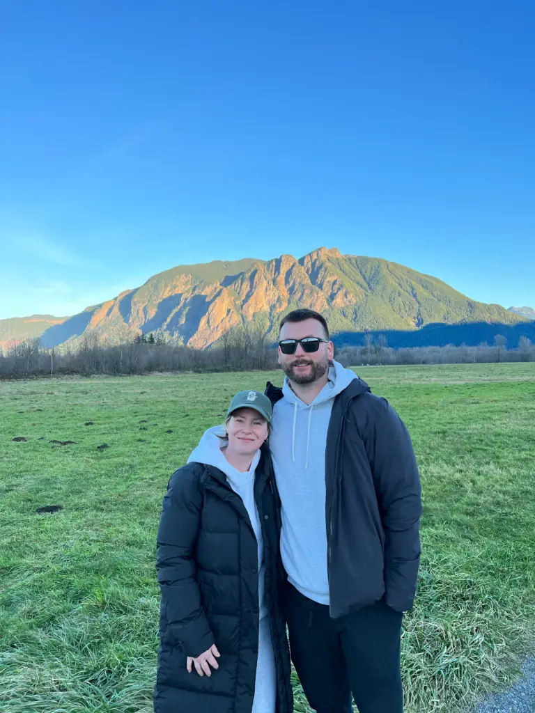 A man in sunglasses and a woman in a green cap standing together on a grassy field with a mountain behind them under a clear blue sky